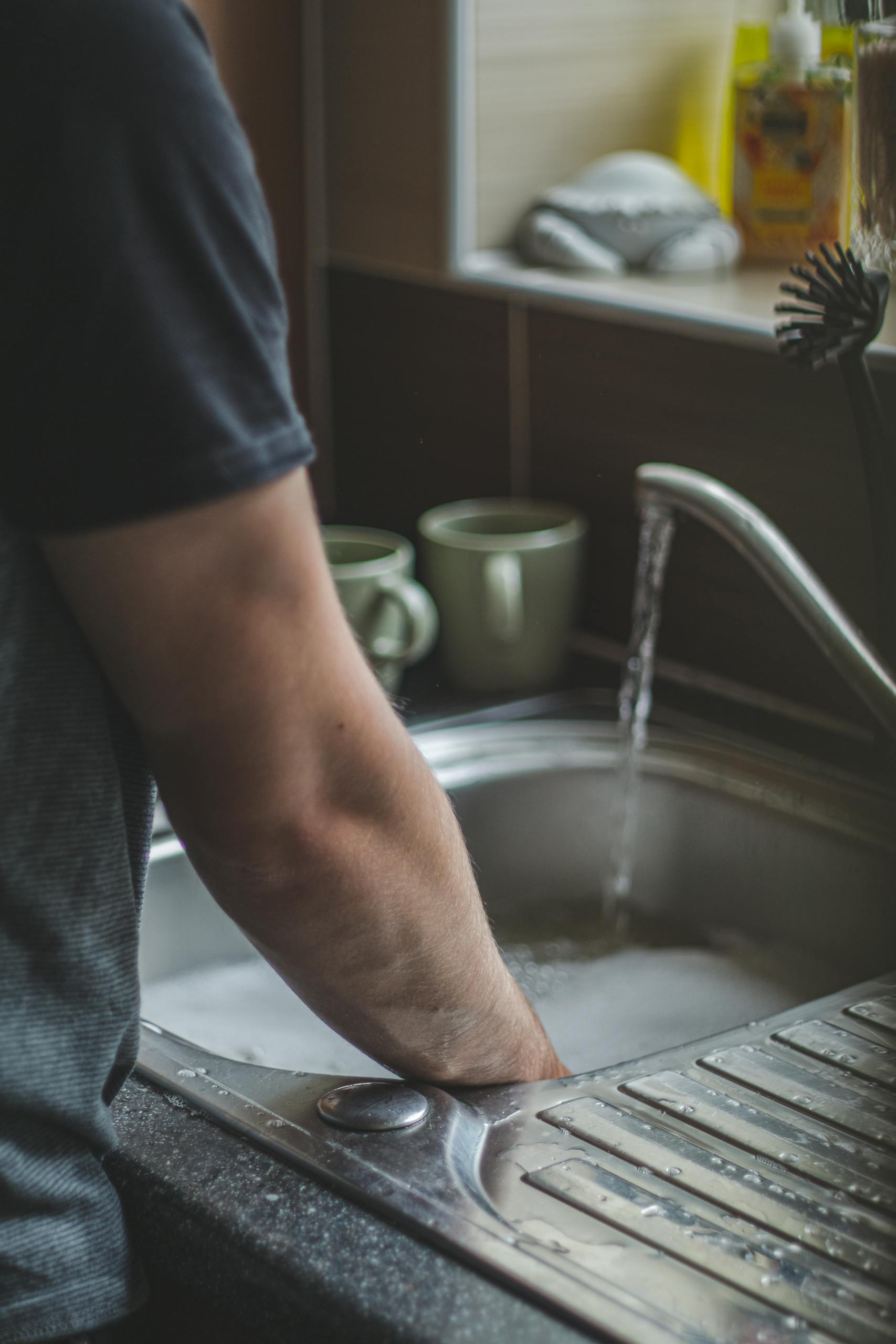 A man washing his hands at a kitchen sink with cups and cleaning products nearby.
